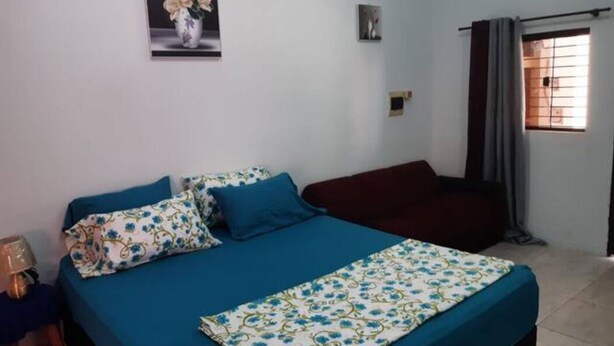 Un dormitorio con cama, almohadas, una colcha, un taburete pequeño, una ventana con cortinas y un interruptor de luz en la pared.