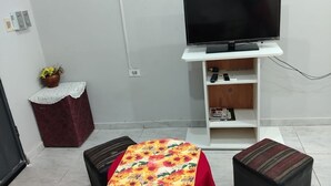 Un pequeño espacio de estar con un televisor sobre un mueble blanco, una mesa con mantel rojo, un banco y un pequeño armario rojo con una planta.