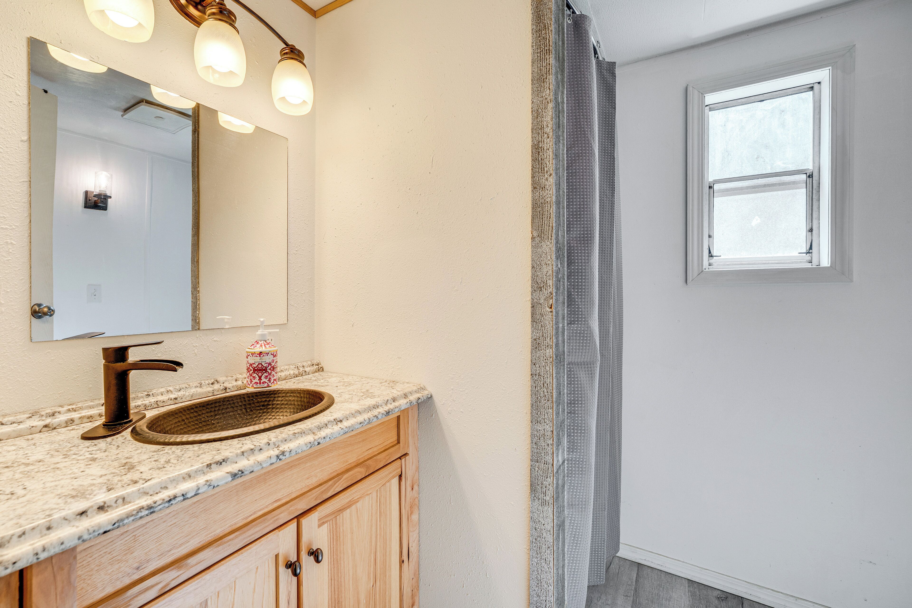 Une salle de bain dotée d’un comptoir en granit, d’un meuble-lavabo en bois, d’un évier, d’un miroir avec éclairage intégré et d’une fenêtre offrant une vue agréable.