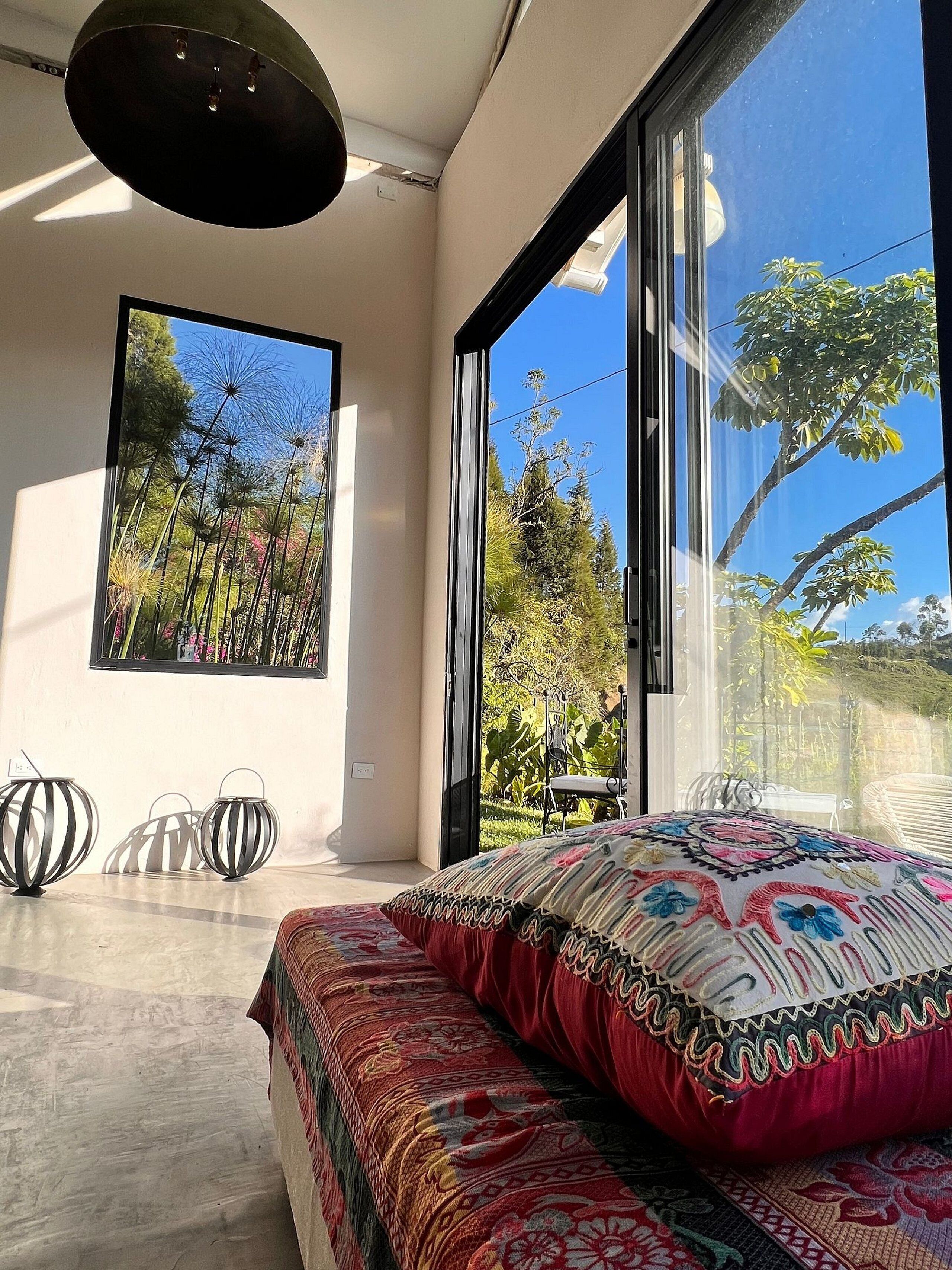 A modern bedroom with a large bed, a framed picture of a garden, and a view of trees through large windows.