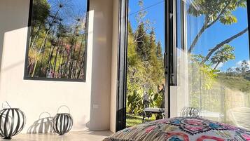 A modern bedroom with a large bed, a framed picture of a garden, and a view of trees through large windows.