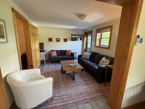 A living room with a sofa, a coffee table, and a white armchair. There are three framed pictures on the wall and a window with a view of greenery.