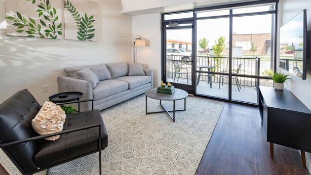 Ein modernes Wohnzimmer mit grauem Sofa, einem schwarzen Ledersessel, einem runden Couchtisch und einem großen Fenster mit Blick auf einen Balkon und umliegende Gebäude.