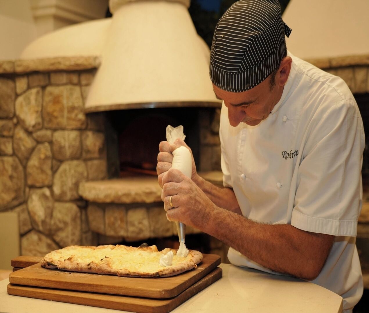 A chef in a white uniform with a striped hat, preparing a pizza on a wooden board in front of a stone oven.