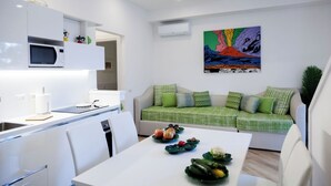 A modern kitchen with white cabinetry, a microwave, and a dining area with a white table and chairs. There is a green sofa with patterned cushions and a colorful abstract painting on the wall. - Resort Lubrense - Fiore Di Campo with shared pool (Sant'Agata Sui Due Golfi)