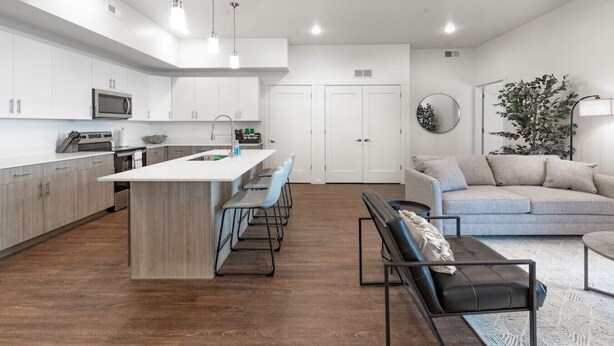 A modern kitchen with white cabinets, a central island, and bar stools. There is a dining area with a sofa and a coffee table. - Apartment 2108 · Luxury style 2 bed 2 bath apartment (North Salt Lake)