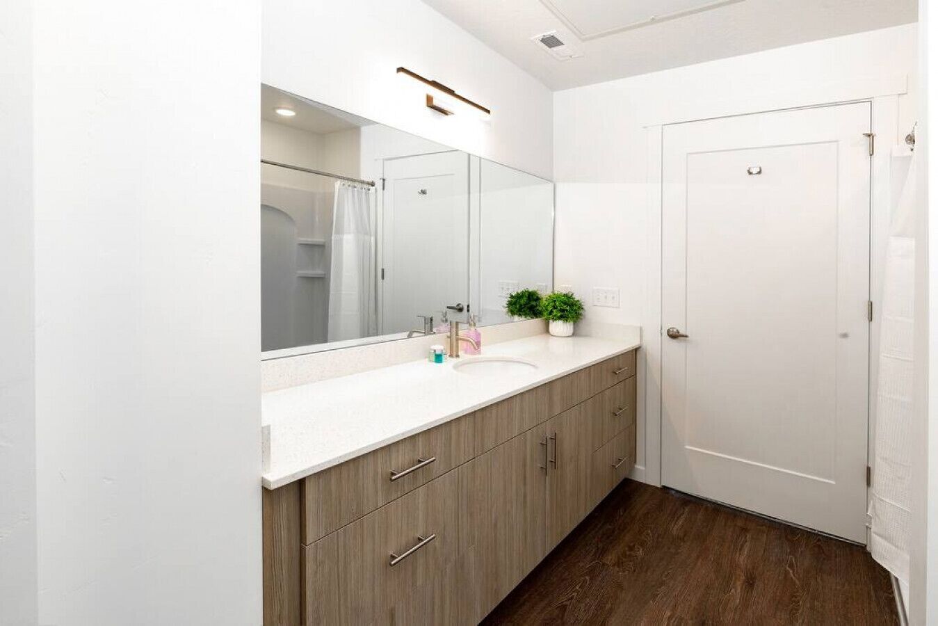 A modern bathroom with a large mirror, a white sink, wooden cabinets, and a door.