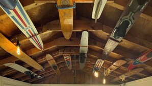 Une pièce avec un plafond haut, des poutres en bois et des planches de surf suspendues aux poutres. Il y a une fenêtre qui laisse entrer la lumière naturelle, un fauteuil en forme de sac noir et une porte qui donne sur l’extérieur.