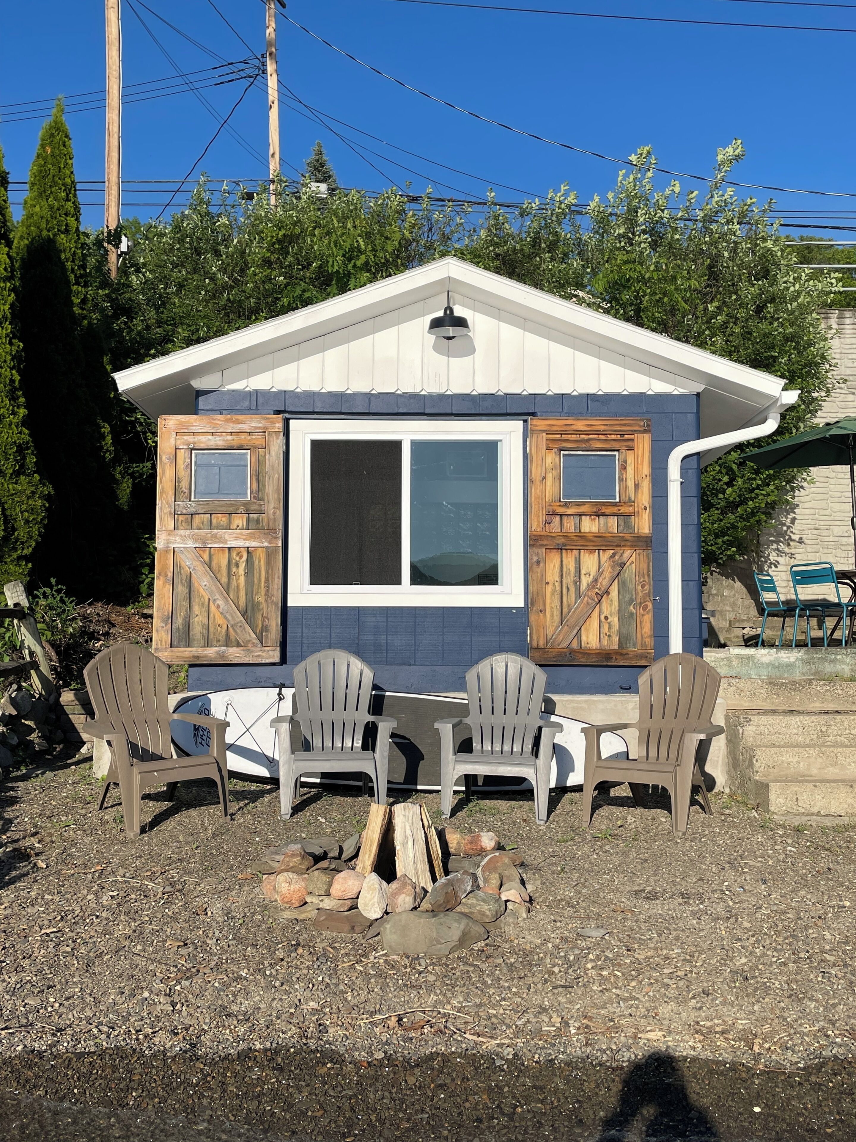 A small cabin with a white and blue exterior, two wooden doors, a large window, and four Adirondack chairs around a central fire pit.