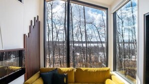 A modern living room with a yellow sofa, a round wooden side table, and a large window offering a view of bare trees.
