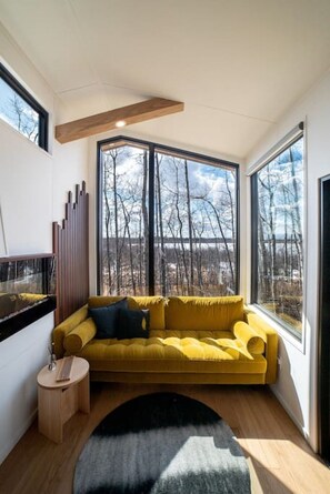 A modern living room with a yellow sofa, a small round wooden side table, and a large window offering a view of bare trees.