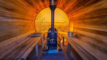 Una sauna con estufa de leña central, bancos y vista a árboles a través de un ventanal circular.