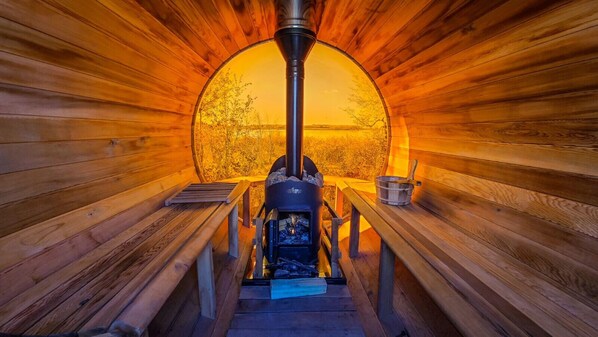 Una sauna con estufa de leña central, bancos y vista a árboles a través de un ventanal circular.
