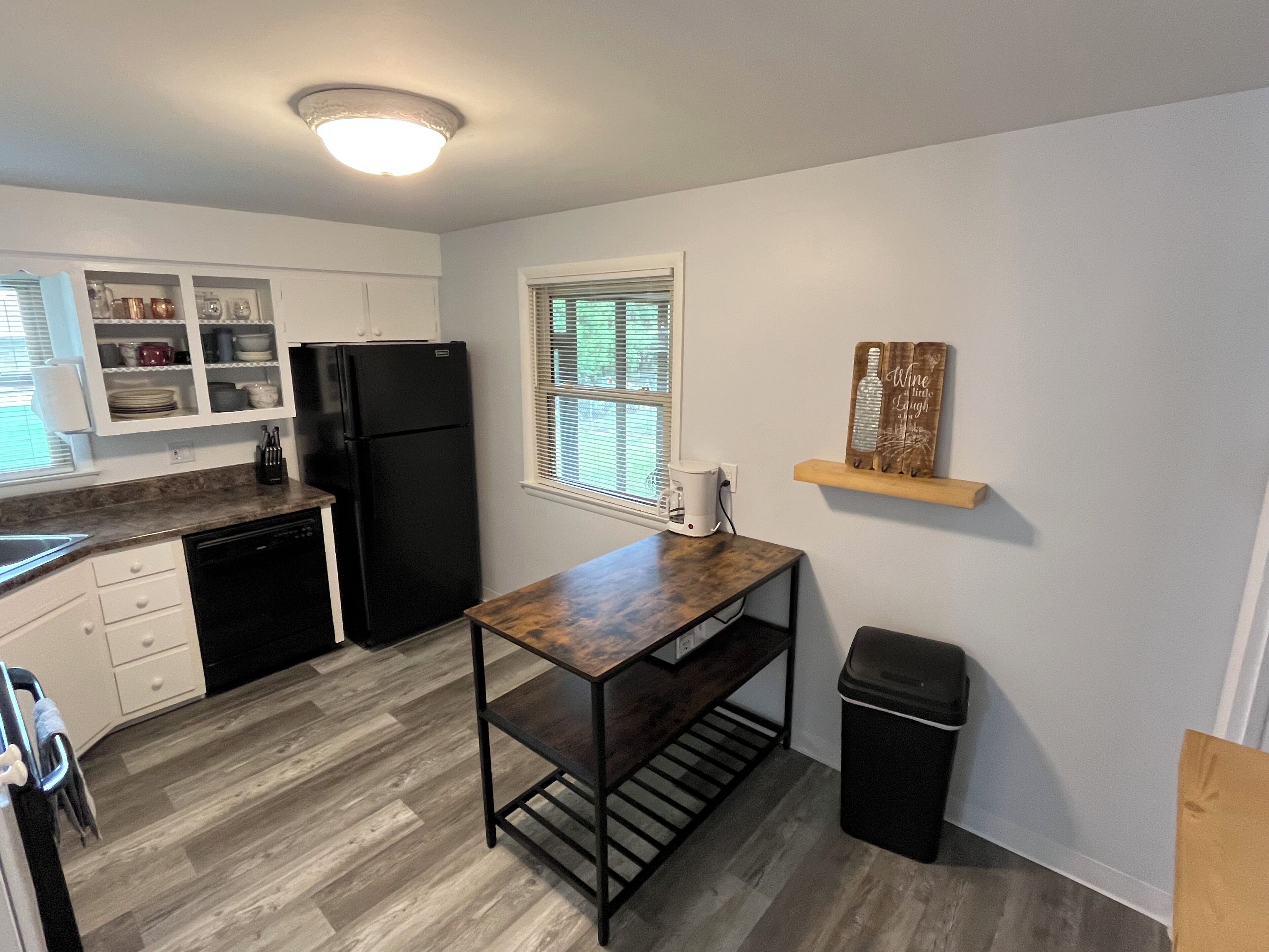 Cocina compacta con armarios blancos, un refrigerador negro, una mesa de madera y una ventana con persianas.