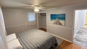 A bedroom with a bed, a ceiling fan, a painting of a beach, and a door leading to another room.