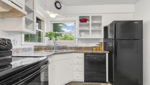 A modern kitchen with white cabinetry, a black refrigerator, a dishwasher, and a window with a view of greenery.