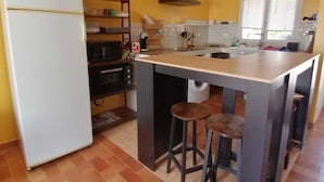 Cocina compacta con refrigerador, microondas, horno y lavamanos. Hay taburetes en la isla y una repisa con utensilios de cocina.
