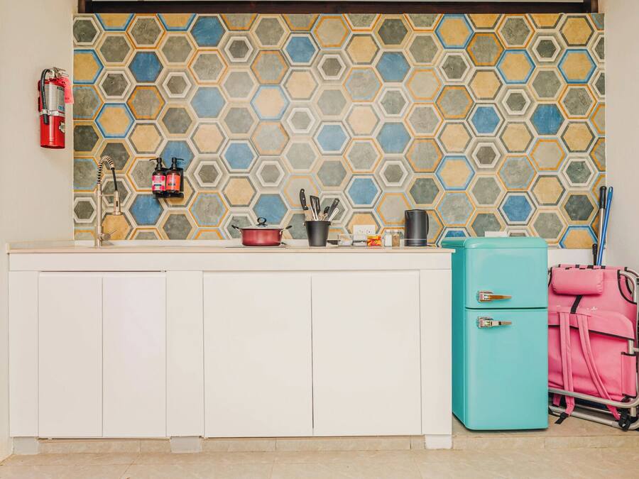 Una cocina con salpicadero de azulejos hexagonales, encimera blanca, maleta rosa y refrigerador verde azulado.