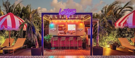 Un área de bar junto a la piscina con un letrero de neón, sillones de descanso y plantas en macetas.