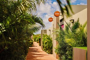 Un camino bordeado de abundante vegetación y palmeras, que conduce a un edificio con letreros numerados E04 y E06.