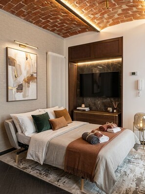 Une chambre moderne avec un plafond en briques, un téléviseur à écran plat, un lit avec de nombreux oreillers, une table de chevet avec une lampe et un radiateur fixé au mur.