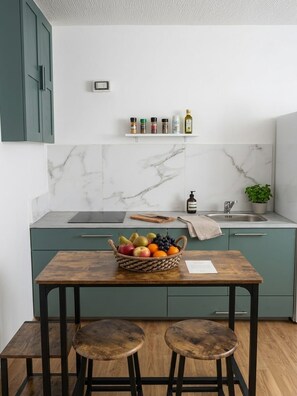 Une cuisine avec un dosseret en marbre, des meubles de couleur turquoise, une table en bois avec des tabourets et un panier de fruits.