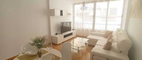 A modern living room with a sofa set, a dining table with chairs, a TV on a white cabinet, and a large window with blinds.