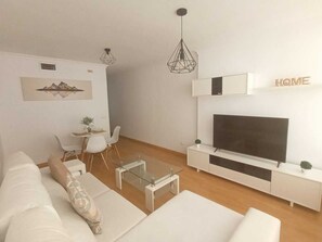 Ein modernes Wohnzimmer mit einem weißen Sofa, einem Glastisch, einer Essecke mit kleinem Tisch und Stühlen, einem Wand-Fernseher und einem Schild mit der Aufschrift 'HOME'.