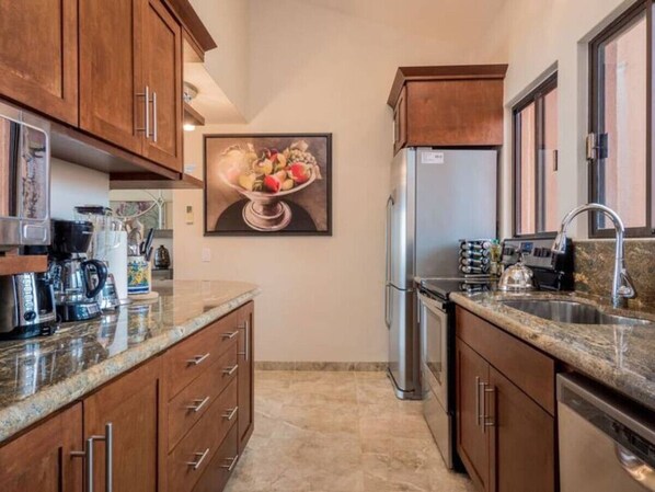 A kitchen with wooden cabinets, a stainless steel refrigerator, a granite countertop, and a painting of fruits and vegetables on the wall.