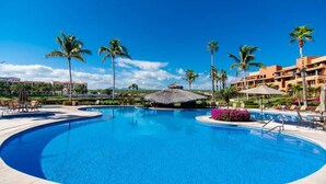 A large, clear swimming pool surrounded by palm trees and lounge chairs, with a thatched-roof structure in the background.