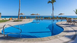 Una piscina con borde curvo, rodeada de sillas de descanso y sombrillas, con vista al océano.