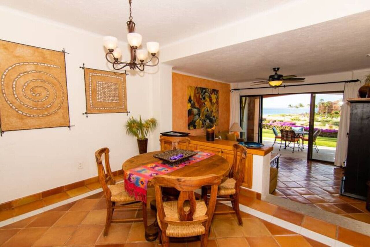 A dining area with a wooden table, chairs, and a chandelier. There is a painting on the wall and a view of the outdoors through a glass door.
