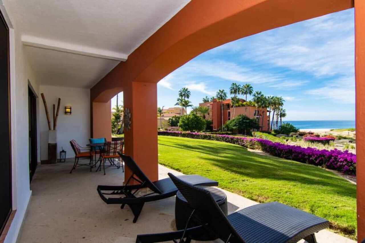 A covered outdoor area with a view of a beach, palm trees, and a well-maintained lawn.