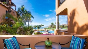 Ein Balkon mit zwei gestreiften Stühlen und einem Tisch, Blick auf einen Pool und eine Resortanlage mit Palmen und blauem Himmel.