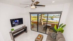 Un salon moderne doté d’un téléviseur à écran plat, d’un ventilateur de plafond, d’un canapé, d’une table basse et d’une porte-fenêtre coulissante donnant sur un espace extérieur avec une piscine et de la verdure.