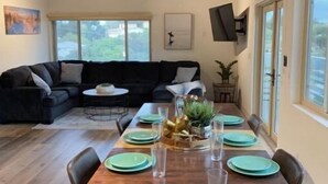 Uma sala de jantar com mesa de madeira posta para quatro pessoas, um sofá preto e um ventilador de teto.