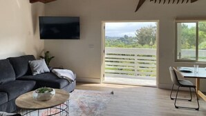 Uma sala de estar moderna com um sofá cinza escuro, uma mesa de centro redonda com uma tigela, teto de madeira, televisão na parede e uma janela grande com vista para o verde.