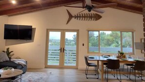 A spacious room with a high ceiling, a ceiling fan, a large fish-shaped light fixture, a flat-screen TV, a dining area with a table and chairs, and a sliding glass door leading to a balcony. - The Medallion Modern City 7bed/5bath 20+ Views/AC (San Diego)