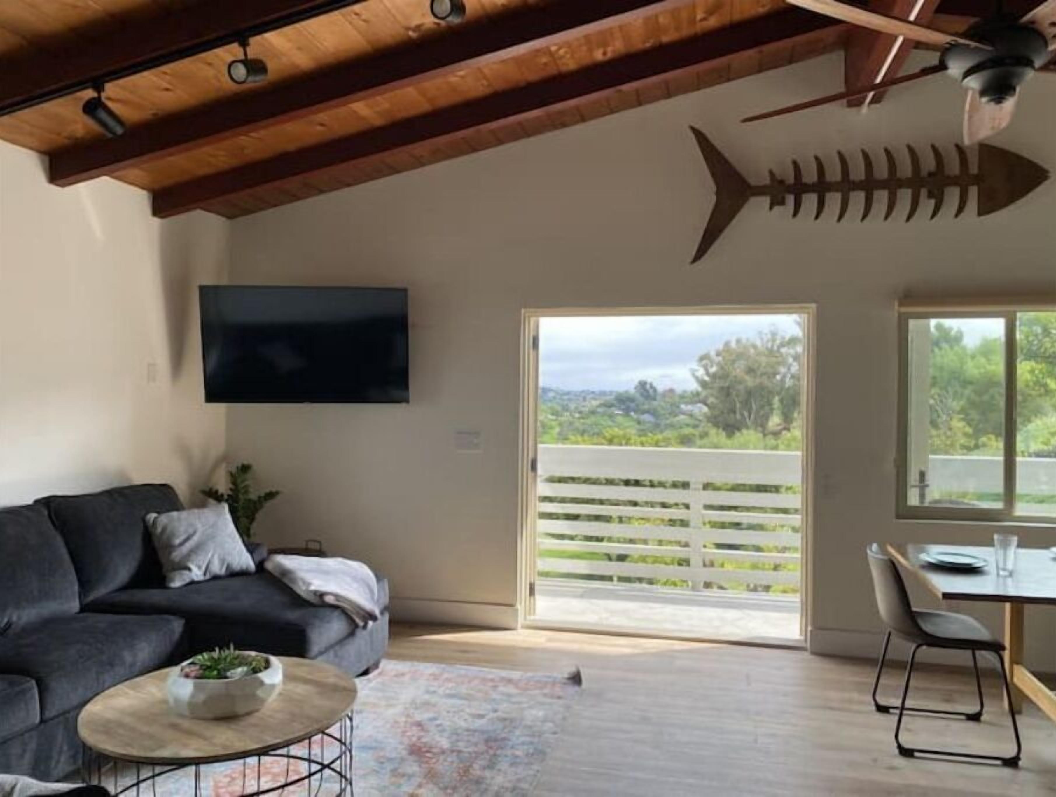 A modern living room with a dark grey sofa, a round coffee table with a bowl, a wooden ceiling, a TV mounted on the wall, and a large window with a view of greenery.