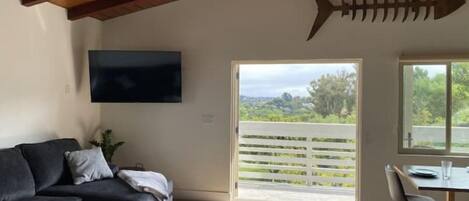 A modern living room with a dark grey sofa, a round coffee table with a bowl, a wooden ceiling, a TV mounted on the wall, and a large window with a view of greenery.