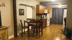 Ein Wohnzimmer mit einem dunkelgrauen Sofa, einem Holzespeisetisch mit Stühlen sowie einer Küchenzeile mit Holzschränken und einer Mikrowelle.