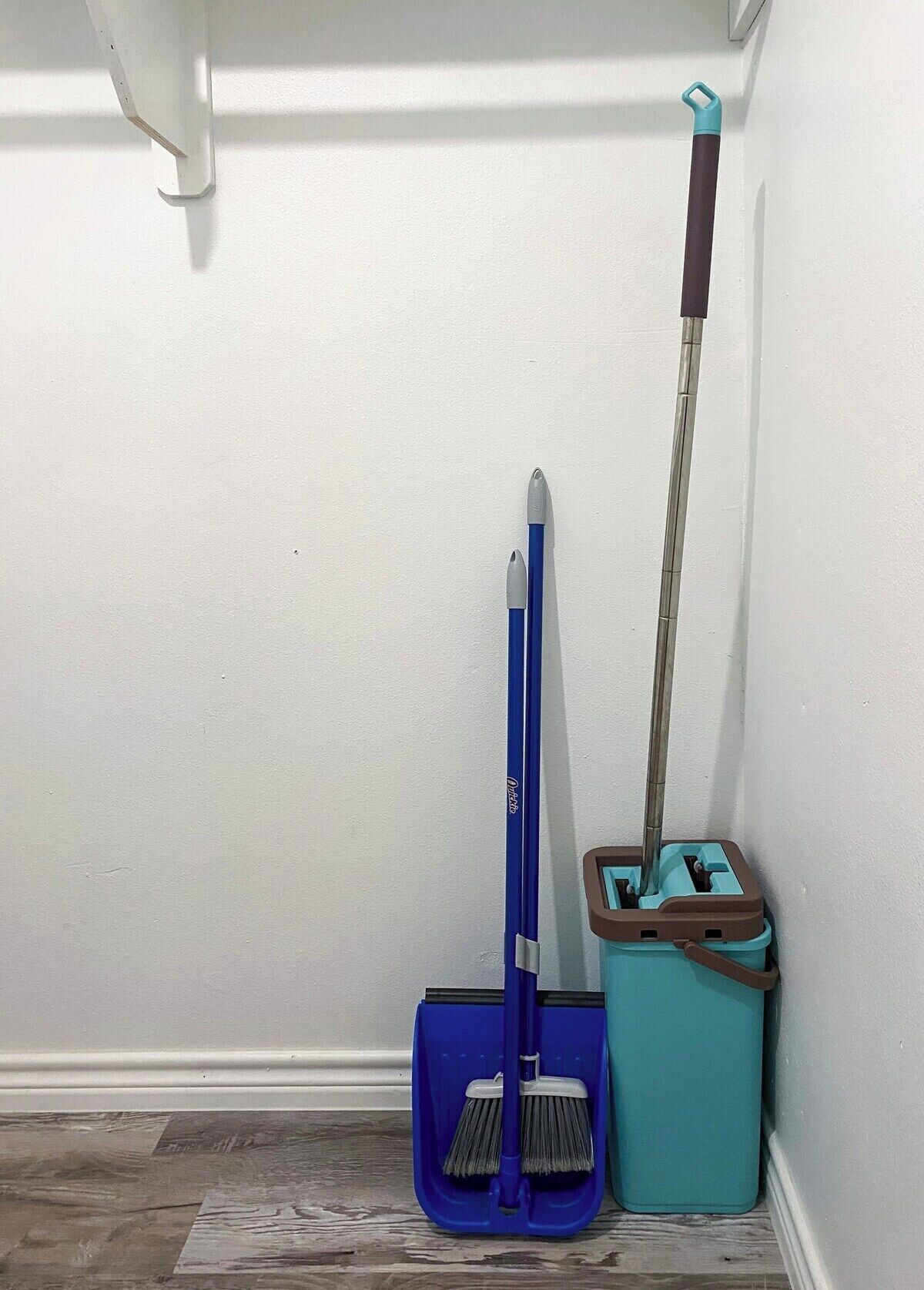 Un rincón de una habitación con una escoba azul y un recogedor turquesa apoyados en la pared.