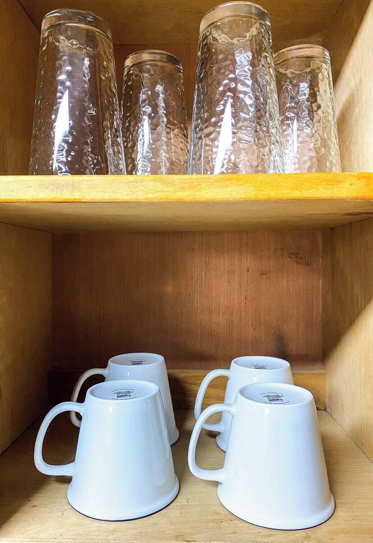 A wooden shelf with four clear glass bottles and four white ceramic mugs.