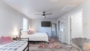 Ein Schlafzimmer mit Bett, Nachttischen, Deckenventilator, an der Wand montiertem Fernseher und einer Tür zu einem anderen Raum.