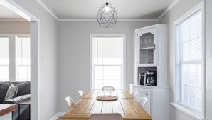 Comedor con mesa de madera, sillas blancas, lámpara colgante y zona de cocina con armarios y cafetera.