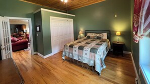 Une chambre avec un plafond en bois, un lit recouvert d’une courtepointe, une table de chevet, une lampe et une porte donnant sur une autre pièce.