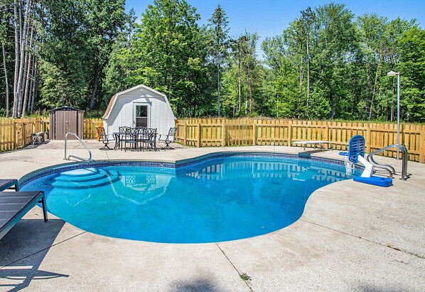 Une piscine avec une terrasse en bois, un petit abri de jardin et une table entourée de chaises.