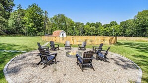 A circular seating area with Adirondack chairs around a fire pit, set on a gravel surface with a wooden fence and trees in the background.