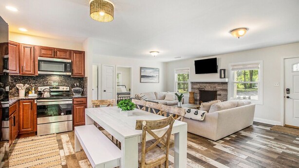 A modern kitchen with wooden cabinets, a built-in microwave, and a large island. There is a dining area with a white table and chairs, and a living room with a sofa and a fireplace.
