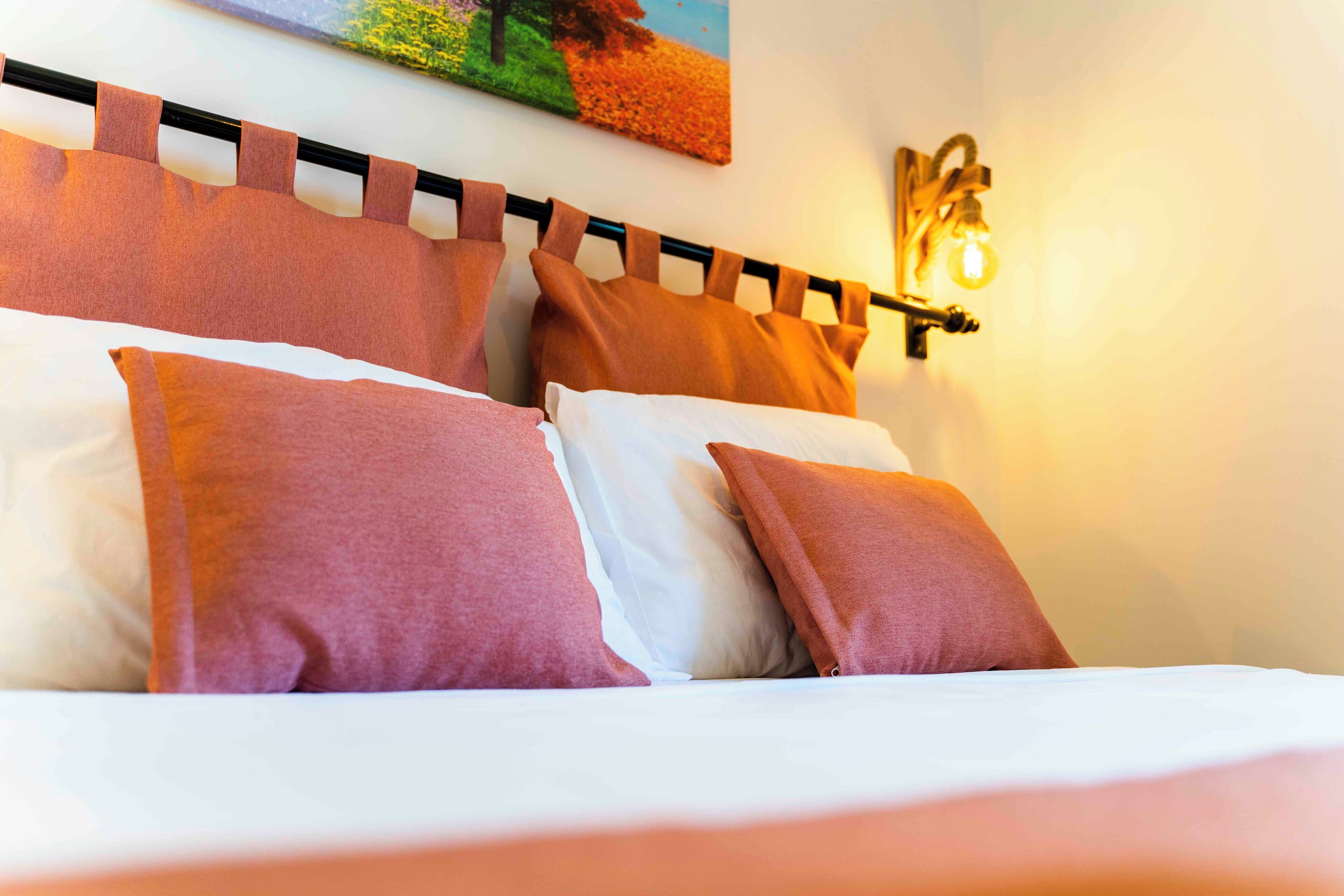 A bed with white and pink bedding, a headboard with a light fixture, and a painting on the wall.